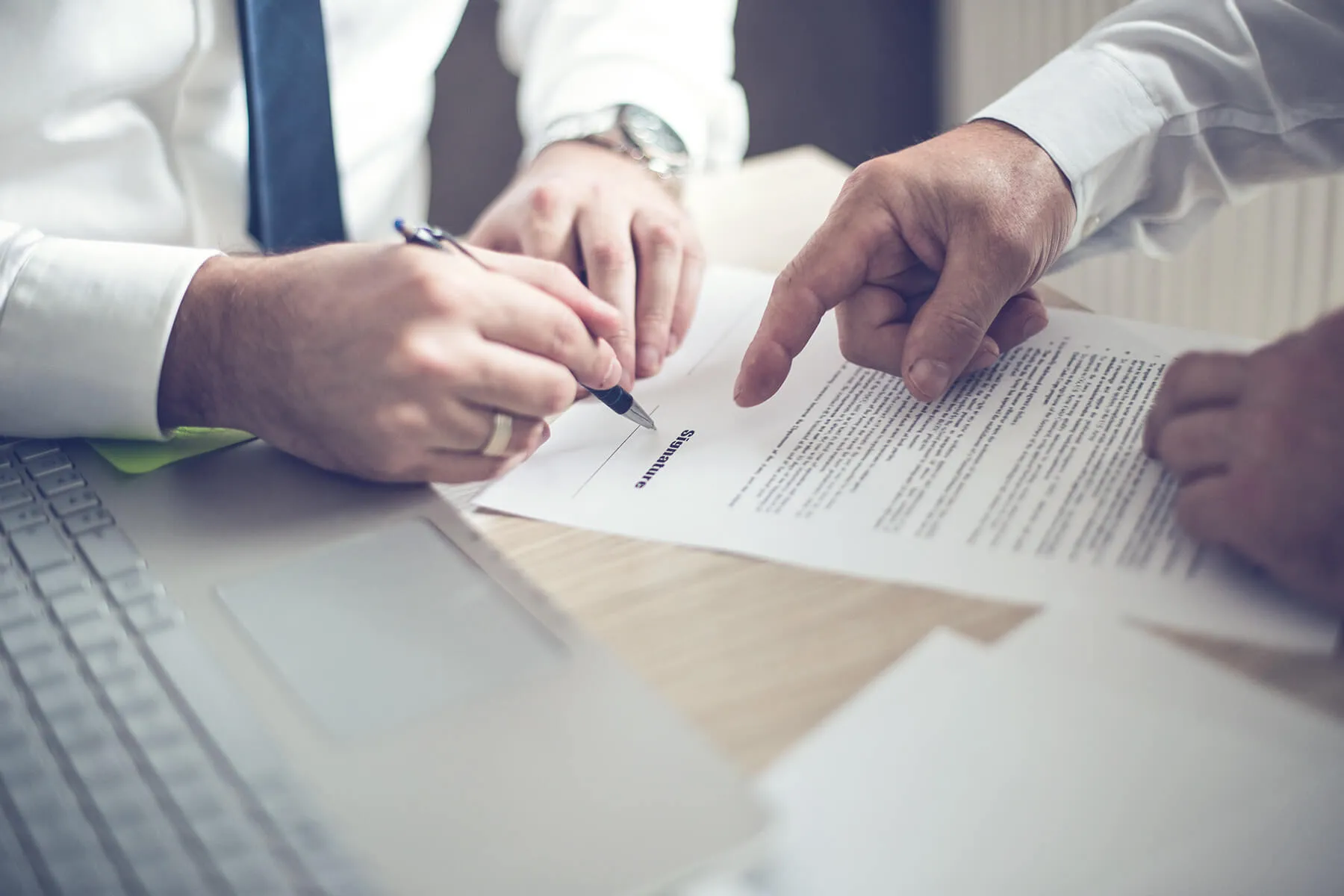 Two individuals signing a legal document over a desk with a laptop and papers.