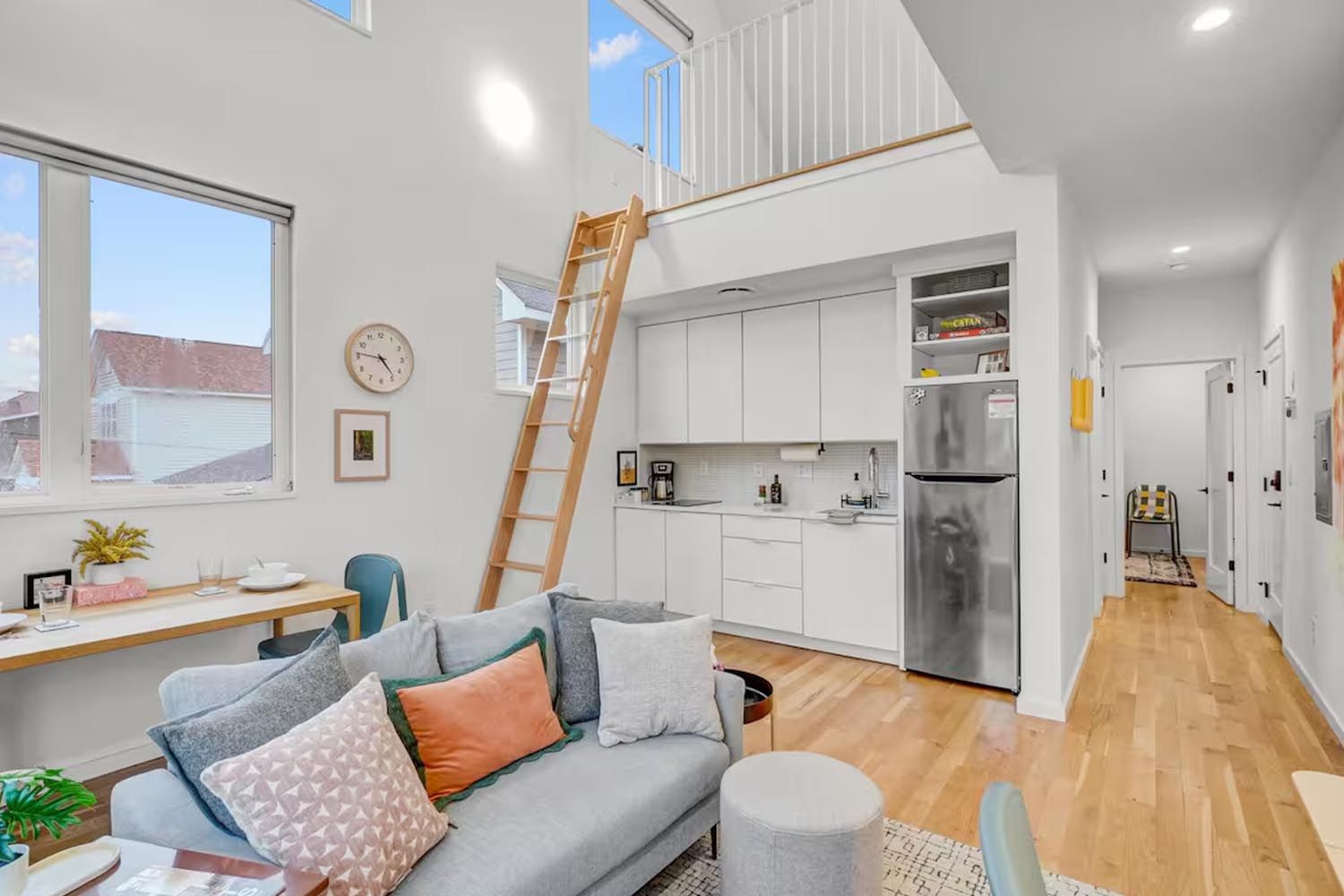 Bright and modern living room with beige sofas, colorful throw pillows, wooden coffee table, black and leather accent chairs, large window with orange curtains, hardwood floors, and decorative wall art.