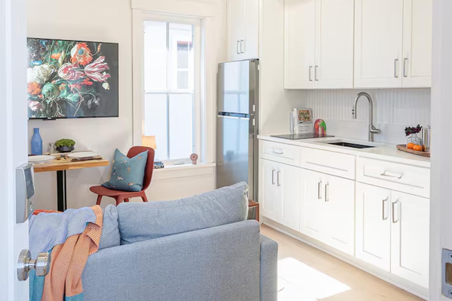 Bright and modern living room with beige sofas, colorful throw pillows, wooden coffee table, black and leather accent chairs, large window with orange curtains, hardwood floors, and decorative wall art.