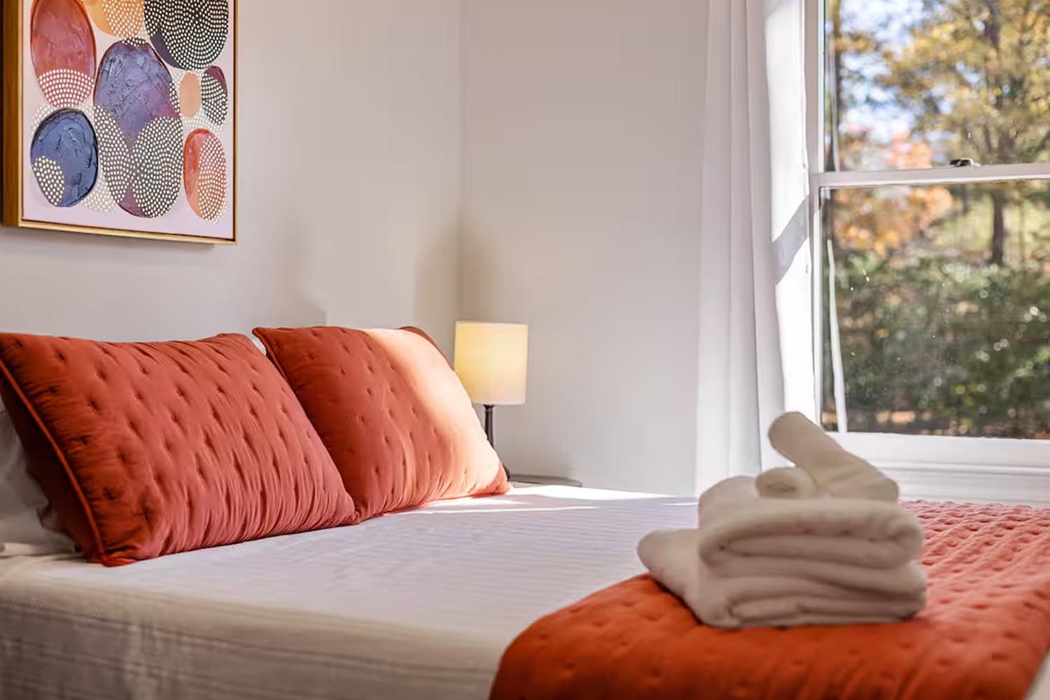 Bright and modern living room with beige sofas, colorful throw pillows, wooden coffee table, black and leather accent chairs, large window with orange curtains, hardwood floors, and decorative wall art.