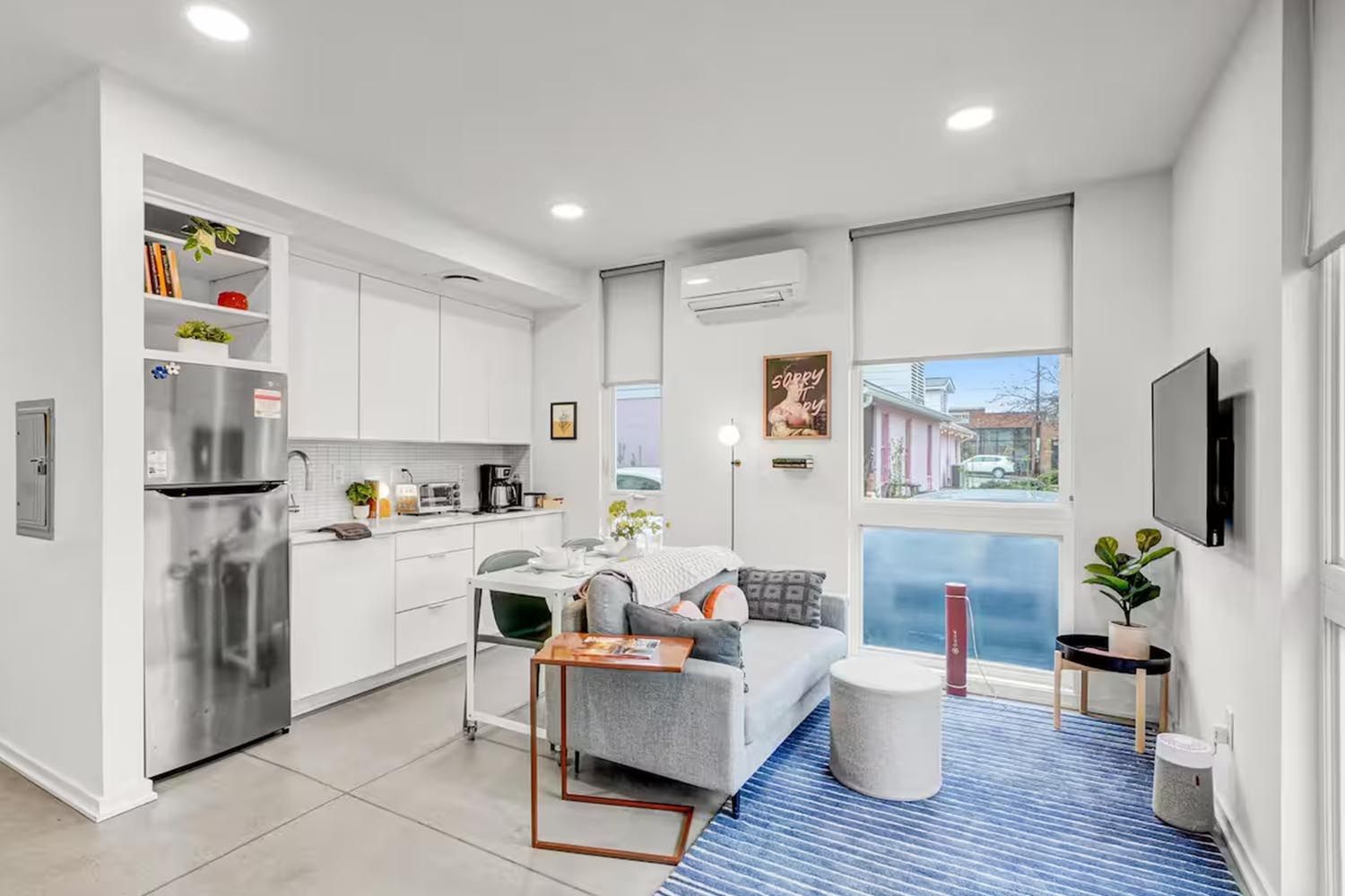 Bright and modern living room with beige sofas, colorful throw pillows, wooden coffee table, black and leather accent chairs, large window with orange curtains, hardwood floors, and decorative wall art.