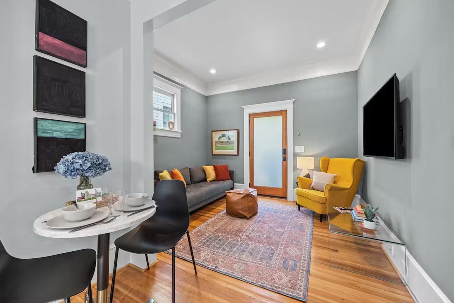 Bright and modern living room with beige sofas, colorful throw pillows, wooden coffee table, black and leather accent chairs, large window with orange curtains, hardwood floors, and decorative wall art.