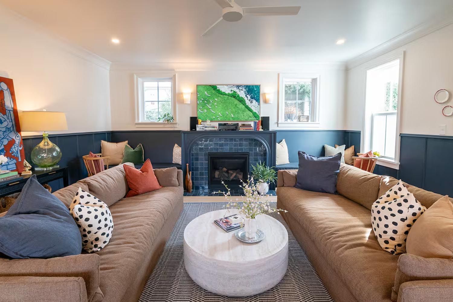Bright and modern living room with beige sofas, colorful throw pillows, wooden coffee table, black and leather accent chairs, large window with orange curtains, hardwood floors, and decorative wall art.