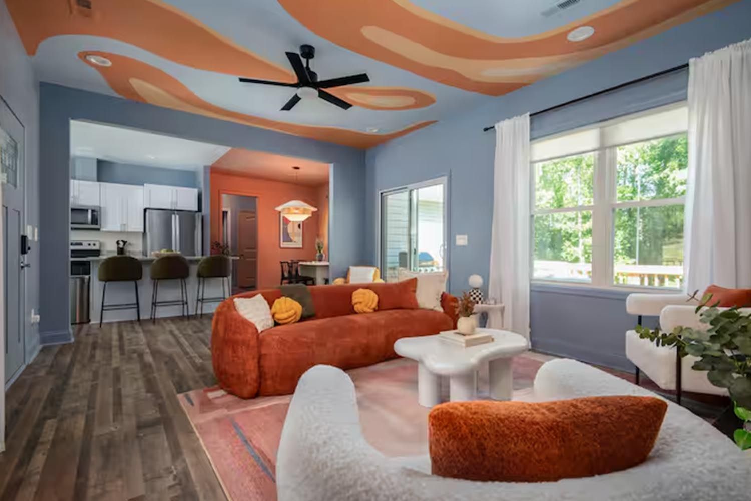Bright and modern living room with beige sofas, colorful throw pillows, wooden coffee table, black and leather accent chairs, large window with orange curtains, hardwood floors, and decorative wall art.