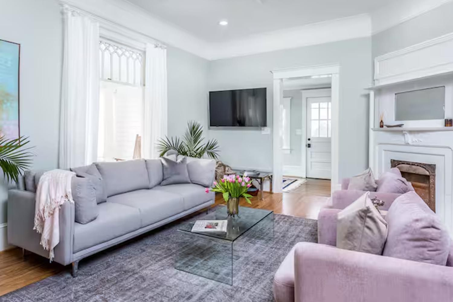 Bright and modern living room with beige sofas, colorful throw pillows, wooden coffee table, black and leather accent chairs, large window with orange curtains, hardwood floors, and decorative wall art.