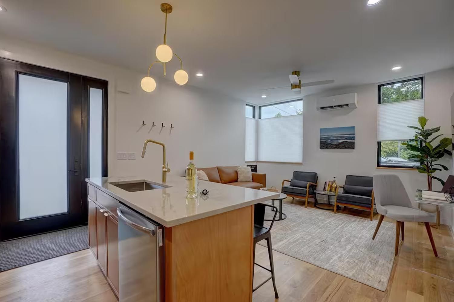 Bright and modern living room with beige sofas, colorful throw pillows, wooden coffee table, black and leather accent chairs, large window with orange curtains, hardwood floors, and decorative wall art.