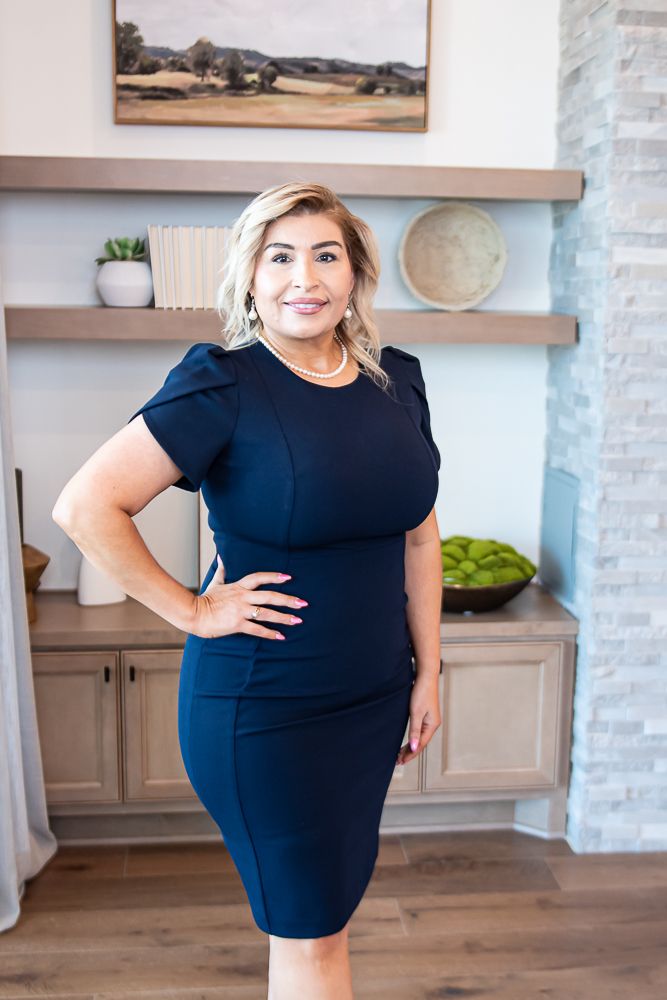 Woman realtor with blonde hair wearing a navy blue dress, standing confidently indoors with a hand on her hip, 
    	        celebrating being recognized as one of the best realtors at TKS.