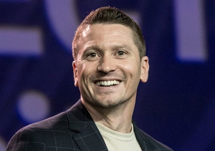 Smiling man in a dark suit jacket and white shirt on stage with blue background.