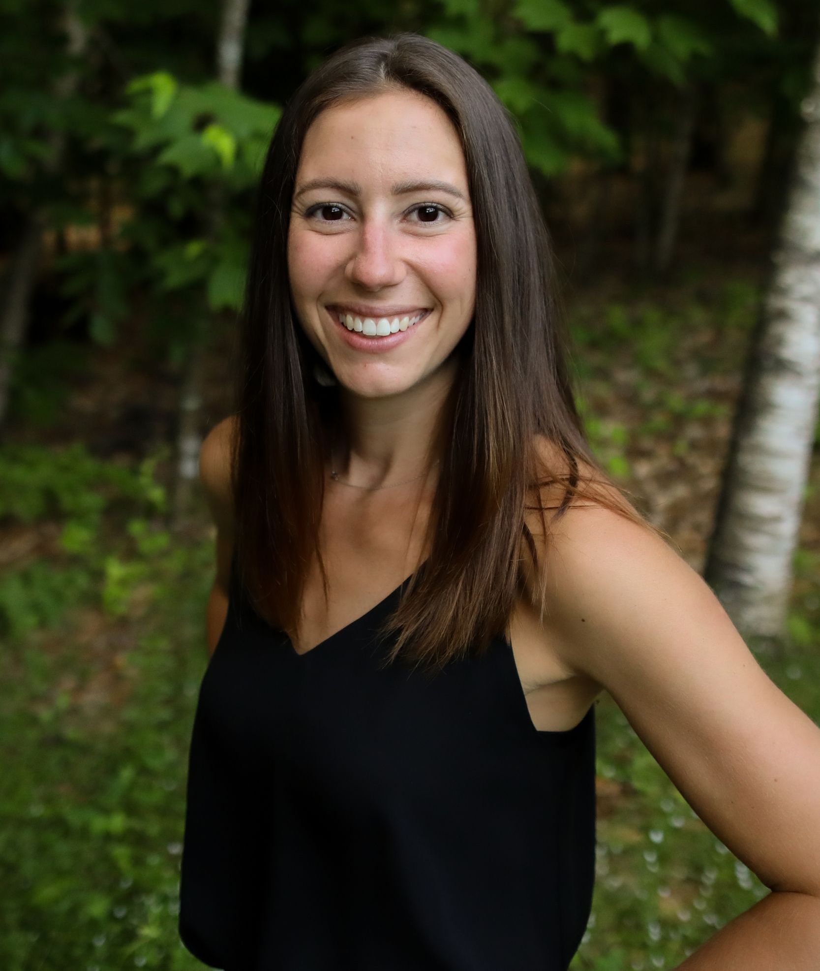 Smile of a woman with long brown hair wearing a black sleeveless top outdoors in a forested area.
