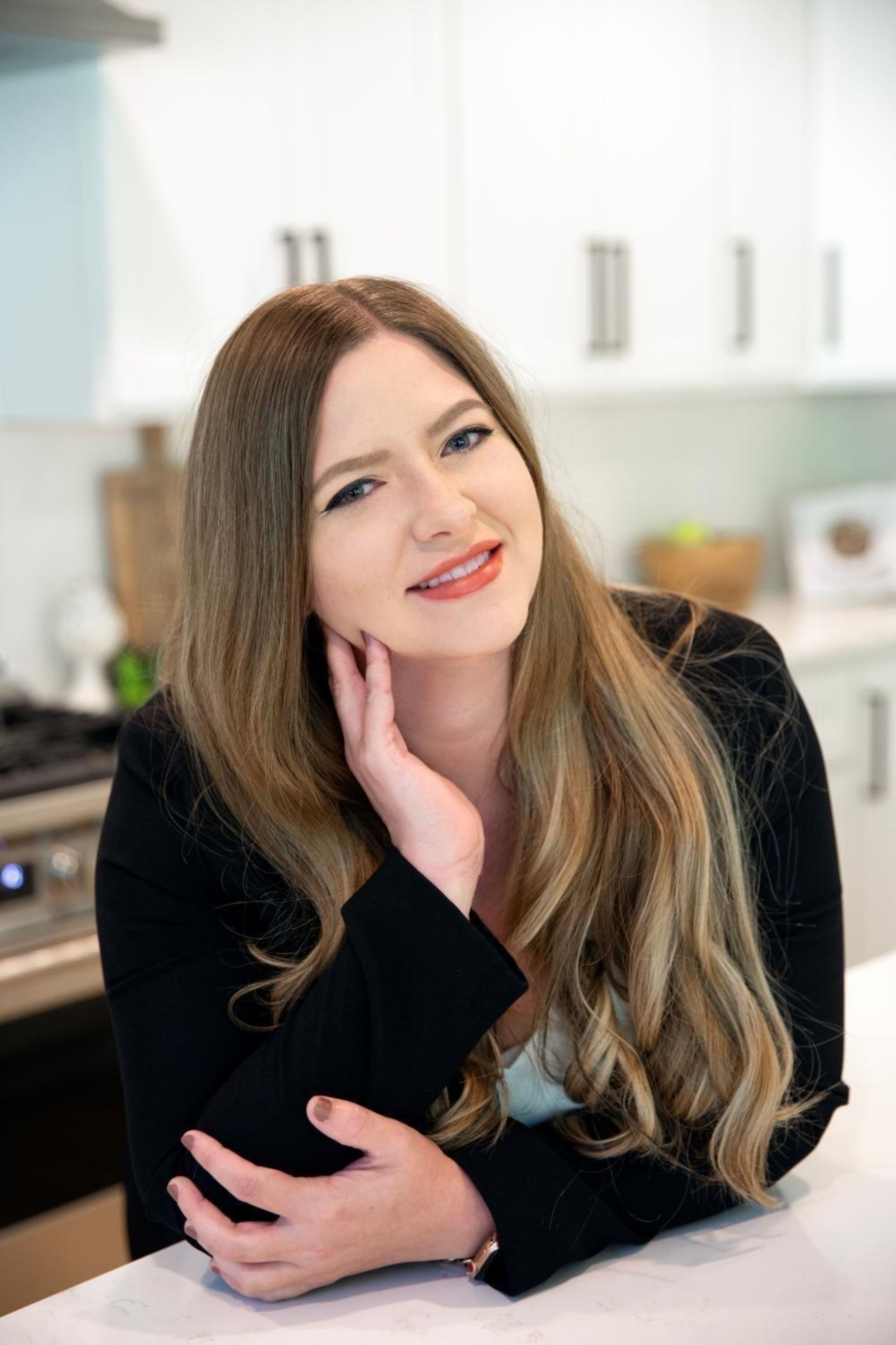 Portrait of a professional woman with long wavy blonde hair, wearing a black blazer and white blouse, standing in an office environment with a friendly smile.