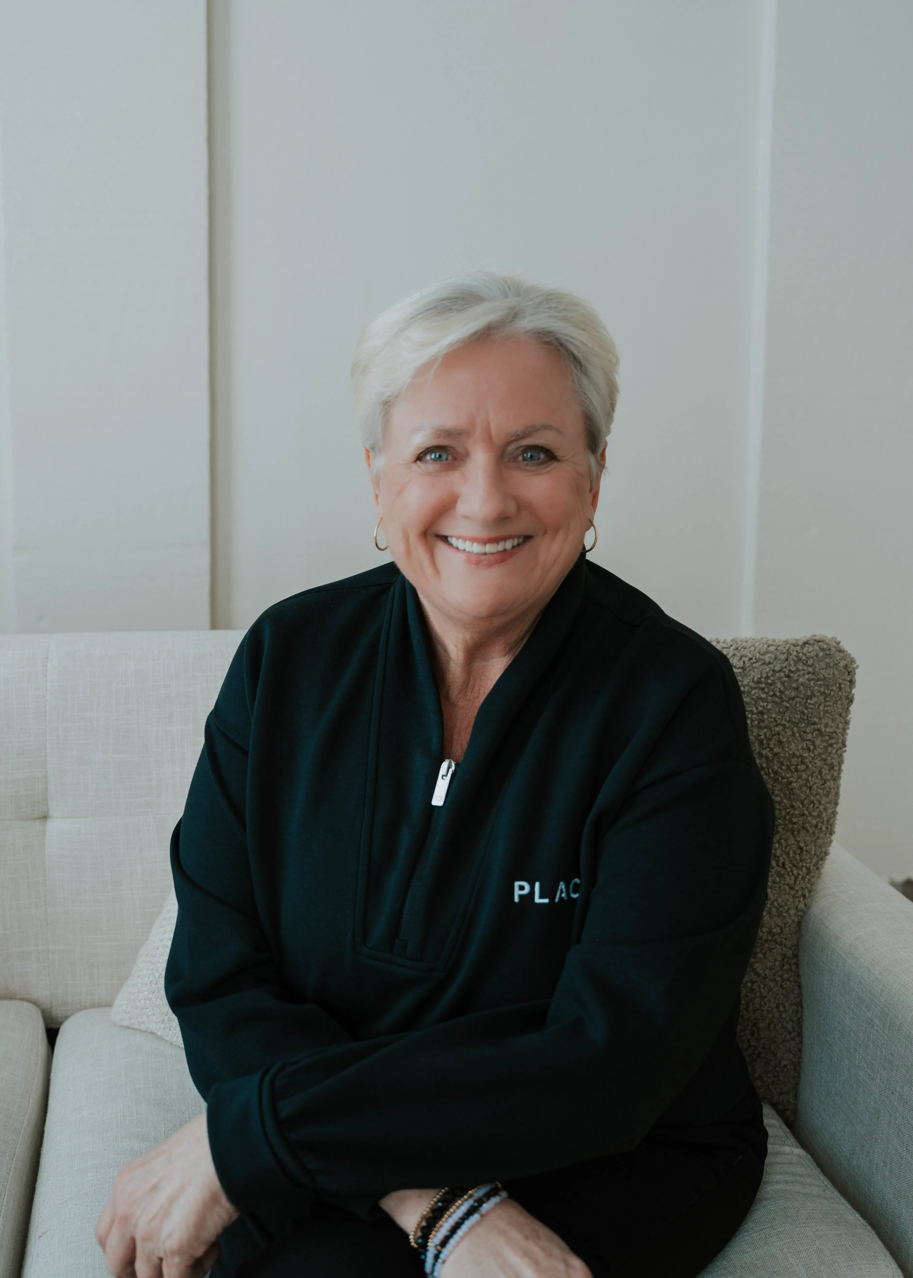Portrait of a smiling middle-aged woman with short blonde hair, wearing gold earrings and a black outfit, against a textured neutral background.