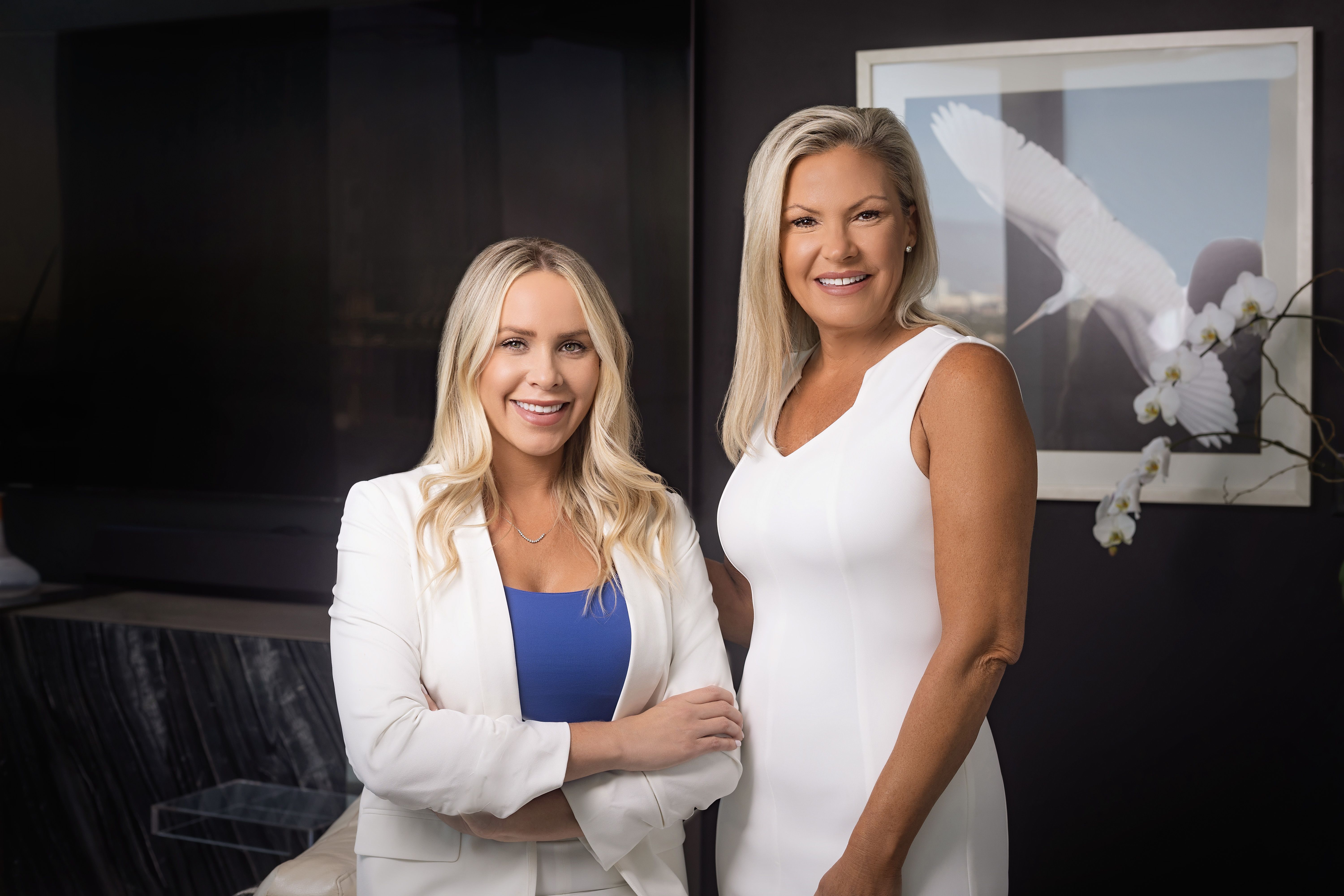 Two smiling women in professional attire standing indoors with a framed artwork of a heron and orchids in the background.