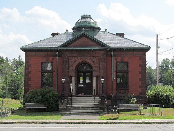 Historic red brick building with a domed roof, front steps, and vintage lampposts, surrounded by greenery and a sidewalk.