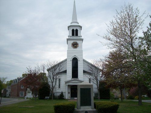 Historic white church with tall steeple and clock tower surrounded by trees and a small monument in the front yard