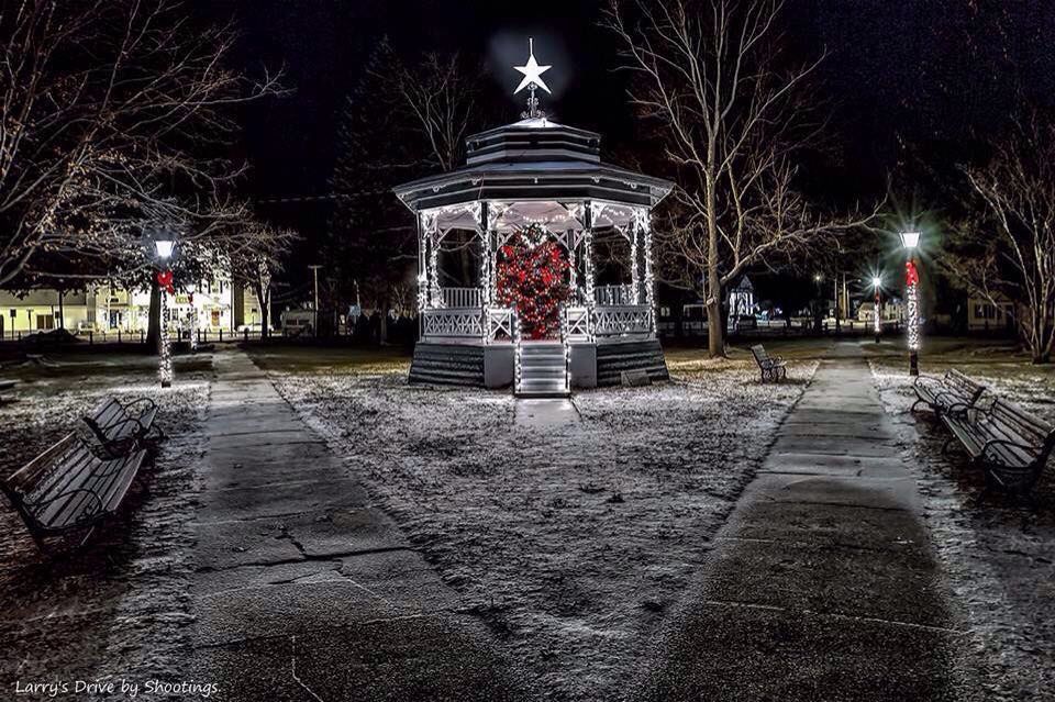 Festive Christmas gazebo decorated with lights and a wreath in a park at night, surrounded by benches and snow, with a large star on top.