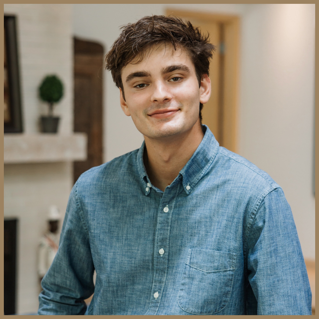 Portrait of a young man with brown hair and a slight smile, wearing a denim shirt indoors.