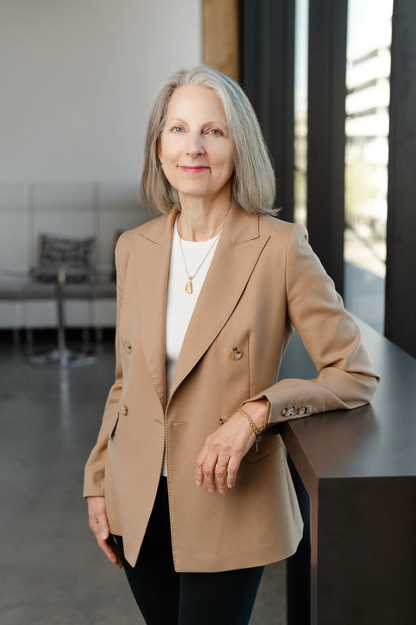Portrait of a mature woman with shoulder-length blonde hair, wearing a white satin blouse and gold jewelry, smiling outdoors with a lush green background.