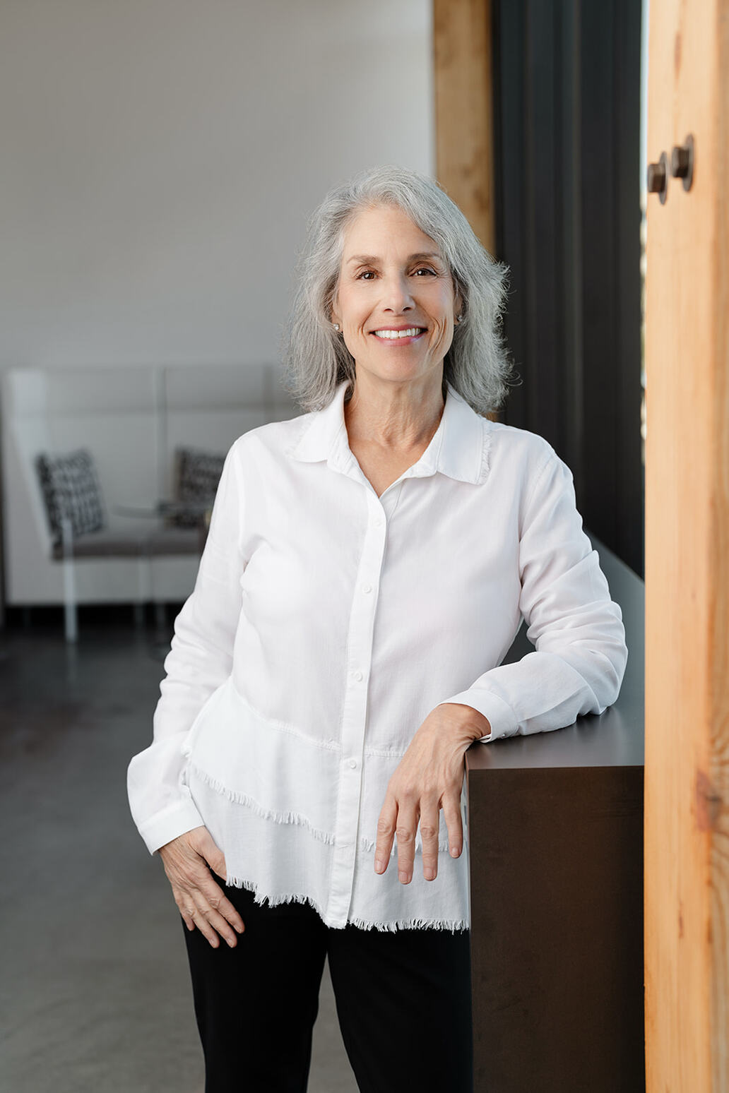 Portrait of a senior woman with gray hair smiling outdoors, wearing pearls jewelry and a navy blue top, with a blurred green garden background.
