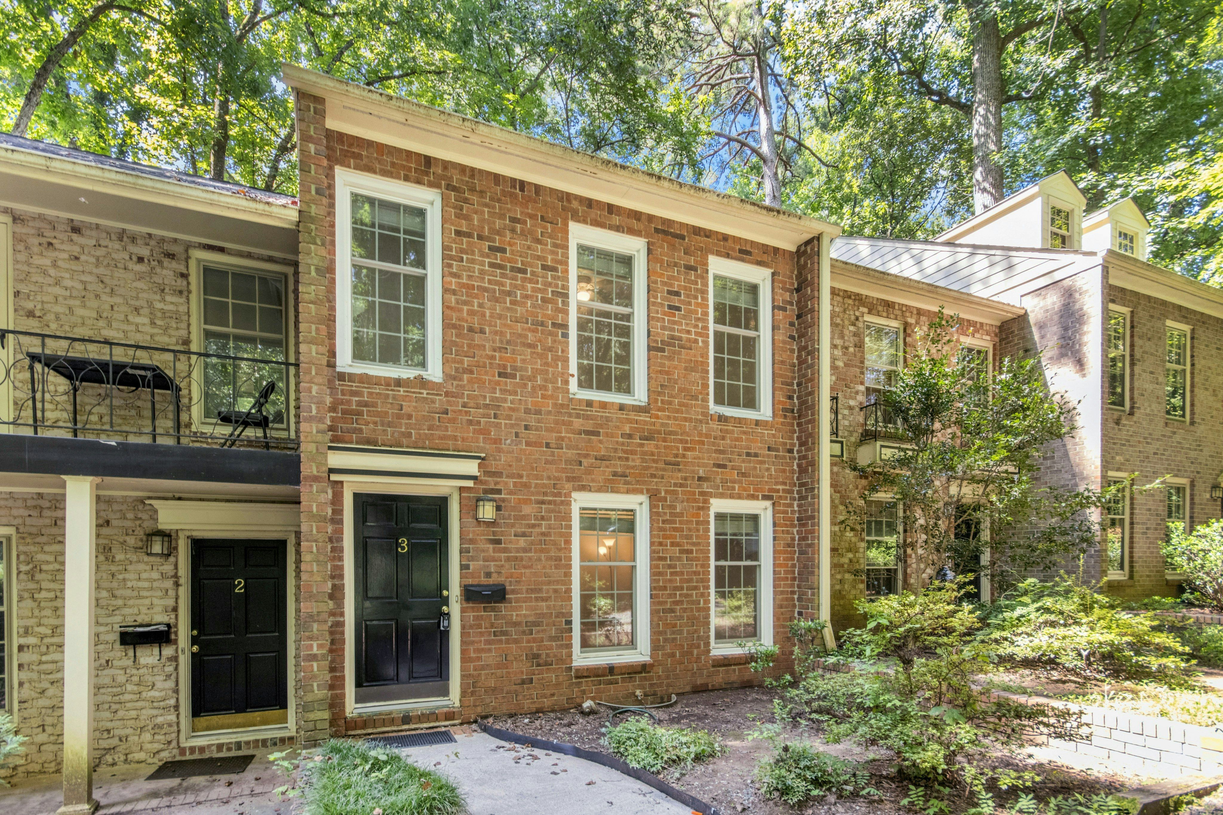 Charming single-story brick home with a well-maintained front yard and lush greenery, surrounded by mature trees under a bright blue sky.
