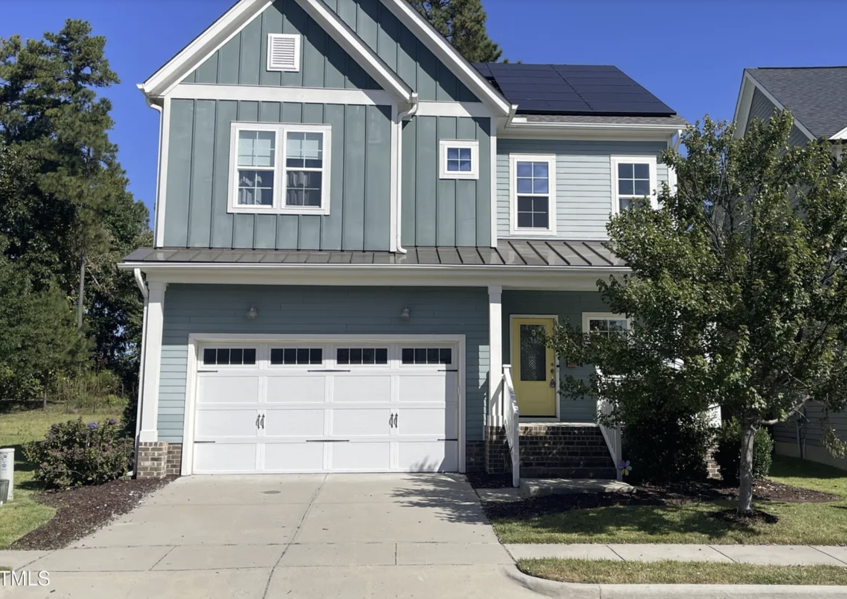 Beautiful new suburban two-story house with white siding, brick accents, dark window shutters, and a front porch with wooden columns, surrounded by manicured lawn and landscaping, featuring a flagpole with an American flag, set against a background of tall trees and a partly cloudy sky.