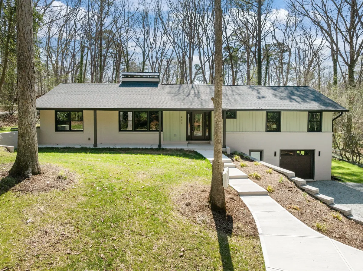 Beautiful new suburban two-story house with white siding, brick accents, dark window shutters, and a front porch with wooden columns, surrounded by manicured lawn and landscaping, featuring a flagpole with an American flag, set against a background of tall trees and a partly cloudy sky.