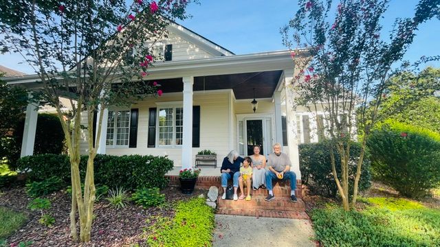 Beautiful new suburban two-story house with white siding, brick accents, dark window shutters, and a front porch with wooden columns, surrounded by manicured lawn and landscaping, featuring a flagpole with an American flag, set against a background of tall trees and a partly cloudy sky.