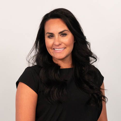Smiling woman with long dark hair wearing a white top standing against a brick wall background