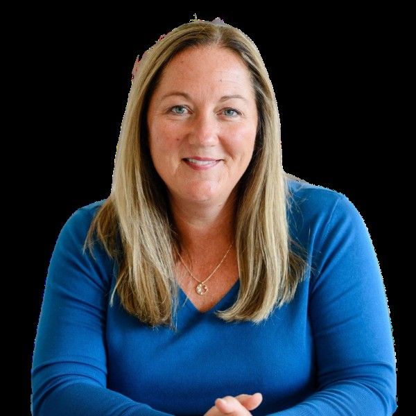 Portrait of a smiling woman with long blonde hair wearing a blue top and a gold necklace, sitting against a black background.