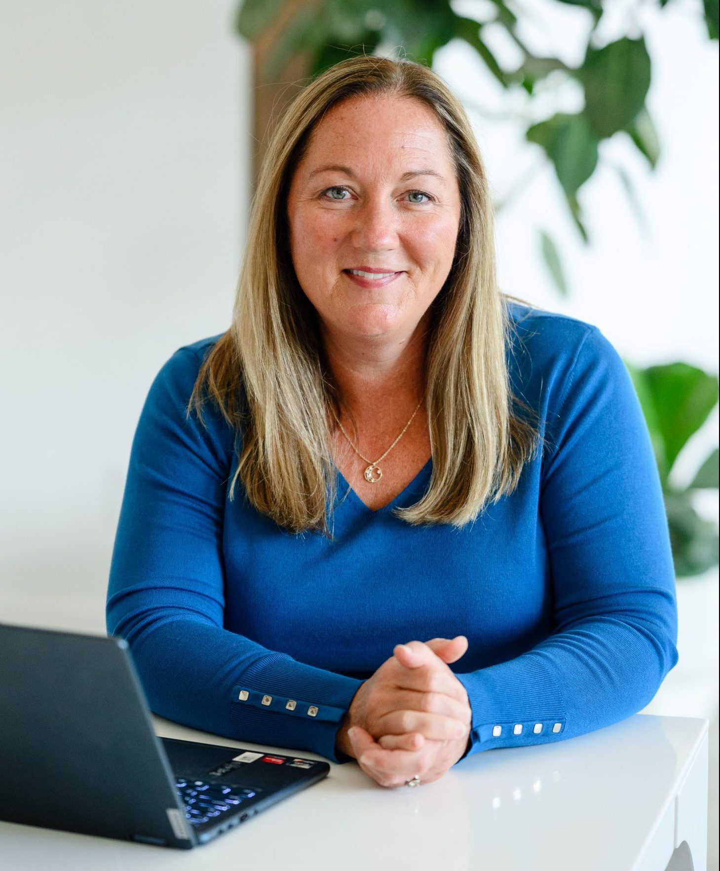 Professional woman smiling at her desk with a laptop and green plants in the background, wearing a blue top and a gold necklace.