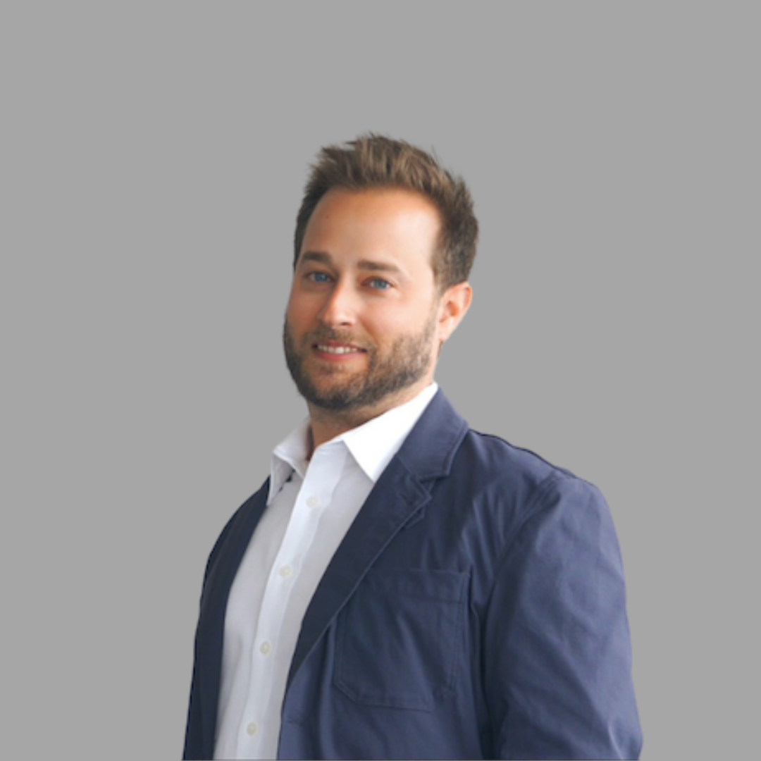 Smiling young man with brown hair and blue eyes outdoors in a green natural setting wearing a white shirt