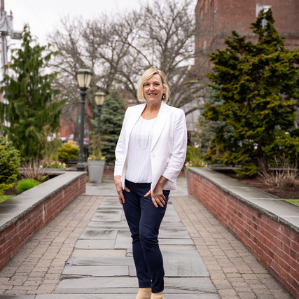 Smiling mature woman in white blazer and navy pants standing on a brick pathway outdoors with trees and street lamps in the background