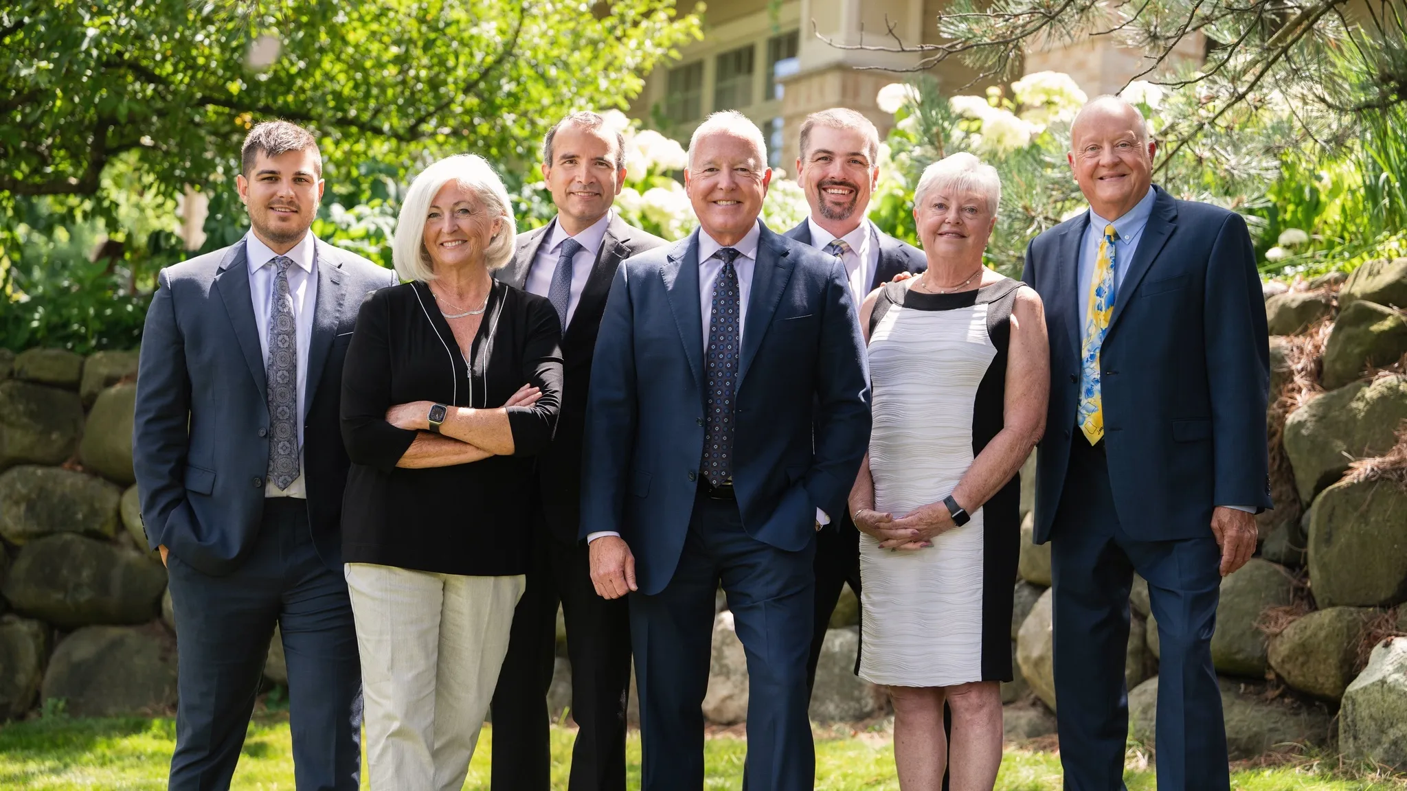 Group of diverse professionals smiling outdoors during daytime, standing in a park with autumn foliage, wearing formal business attire.