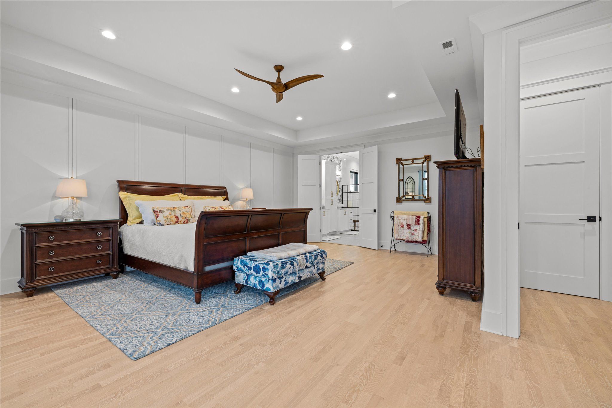 Spacious bedroom with wooden furniture, white paneled walls, hardwood flooring, and modern lighting fixtures.